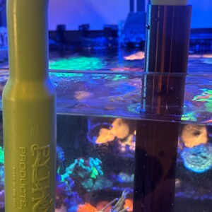 May include: A green plastic aquarium siphon with a dark brown intake tube submerged in water. The siphon has the text "EZ-ON PRODUCTS, INC." printed on it. The aquarium water is filled with colourful coral and plants.