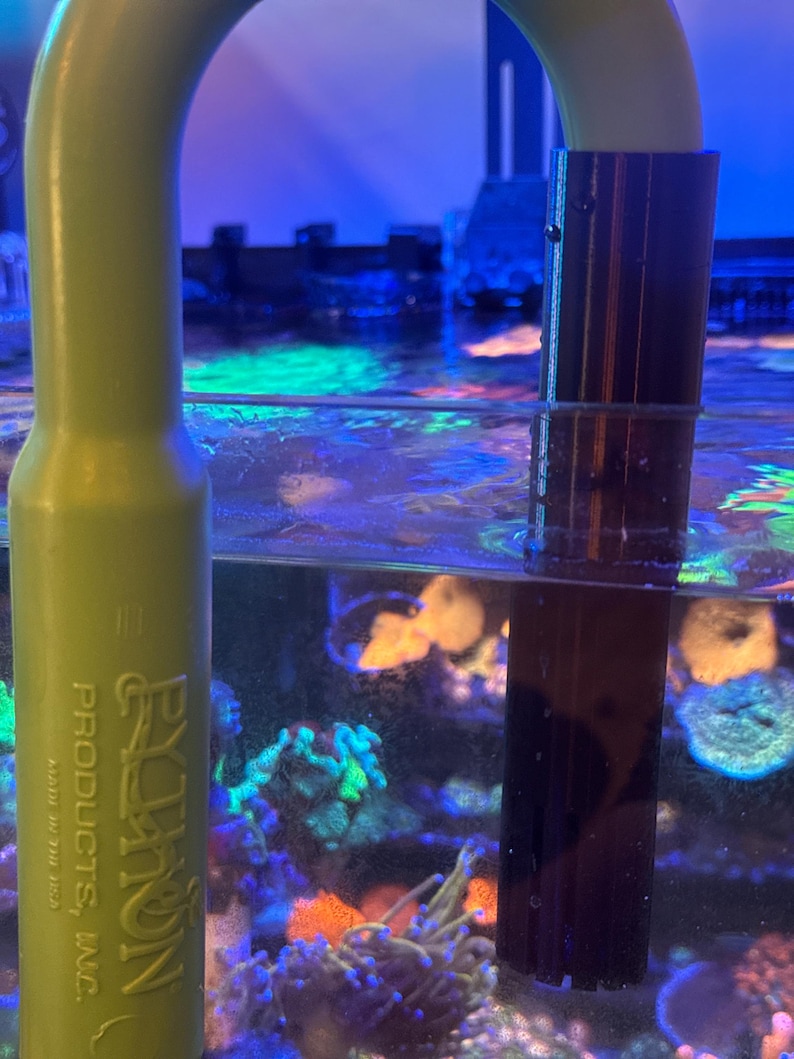 May include: A green plastic aquarium siphon with a dark brown intake tube submerged in water. The siphon has the text "EZ-ON PRODUCTS, INC." printed on it. The aquarium water is filled with colourful coral and plants.