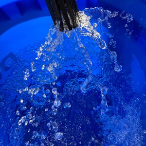 May include: A close-up shot shows water cascading from a black spout into a vibrant blue bucket. The clear water forms droplets and bubbles, contrasting with the blue surface. The image highlights the water's movement and texture.