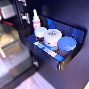 May include: A black, wall-mounted shelf holding various small containers. The containers include a white jar with a QR code, a blue-lidded jar, a white jar with a black top, and a small bottle with a pink label. A blue and white test strip is also visible.