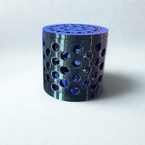 Könnte beinhalten: Ein zylindrischer Behälter in Schwarz mit einem perforierten Design. Der Körper weist zahlreiche kreisförmige Öffnungen auf, die ein blaues Inneres freilegen. Die Oberseite ist ebenfalls blau und hat viele kleine Löcher. Der Artikel ist wahrscheinlich 3D-gedruckt.