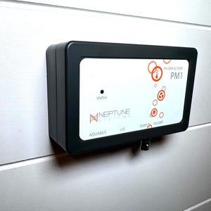 May include: A black rectangular Neptune Systems PM1 device mounted on a white wall. The device has a white face with orange graphics and text, including "pH/ORP & TEMP" and "PM1". The word "status" is visible.