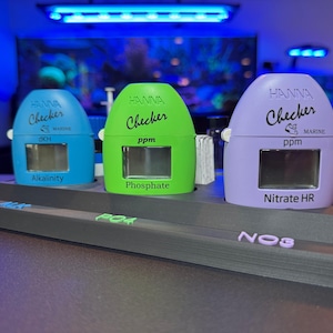 May include: Three Hanna Checker marine water testing devices in blue, green, and purple, each with a digital display. The blue checker reads "Alkalinity", the green reads "Phosphate", and the purple reads "Nitrate HR". They sit on a dark gray stand.