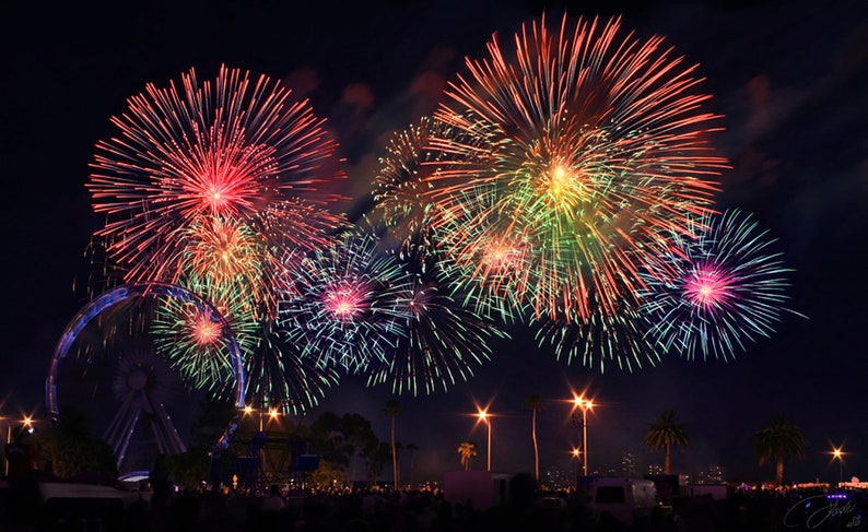 May include: A night sky filled with colorful fireworks exploding over a Ferris wheel and a crowd of people. The fireworks are red, green, blue, and yellow.