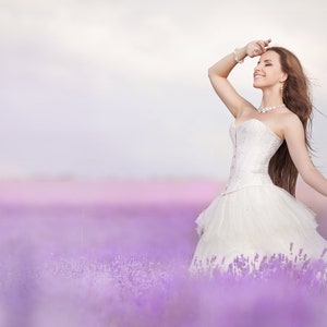 Puede incluir: Una novia con un vestido de novia blanco con un corpi&ntilde;o de encaje se encuentra en un campo de flores de lavanda. Sonr&iacute;e y sostiene un ramo de lavanda.