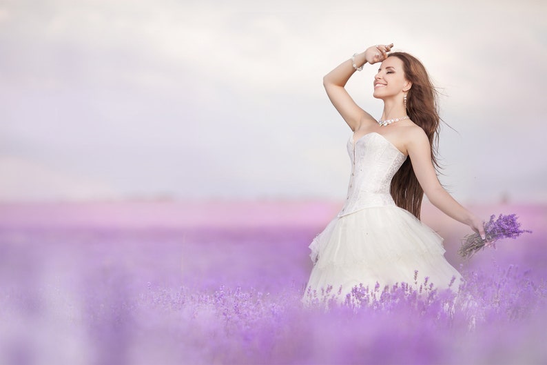 Puede incluir: Una novia con un vestido de novia blanco con un corpi&ntilde;o de encaje se encuentra en un campo de flores de lavanda. Sonr&iacute;e y sostiene un ramo de lavanda.