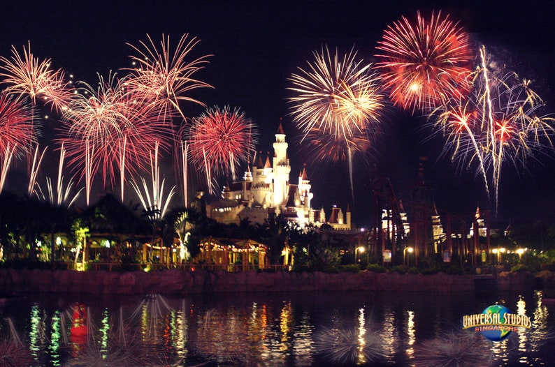 May include: A nighttime view of a castle-like building with fireworks exploding over the water in front of it. The fireworks are red, white, and blue. The text "Universal Studios Singapore" is visible in the bottom right corner of the image.