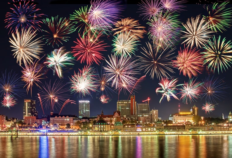 May include: A nighttime cityscape with a vibrant fireworks display. The sky is filled with bursts of red, green, yellow, and purple fireworks. The city skyline is illuminated with lights reflecting on the water below.