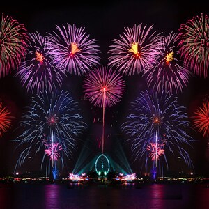 May include: A night sky filled with colorful fireworks exploding over a city skyline. The fireworks are a mix of red, pink, blue, and white.