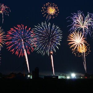 May include: A night sky filled with colorful fireworks exploding in a variety of shapes and sizes. The fireworks are set off in a field with a few buildings in the background.