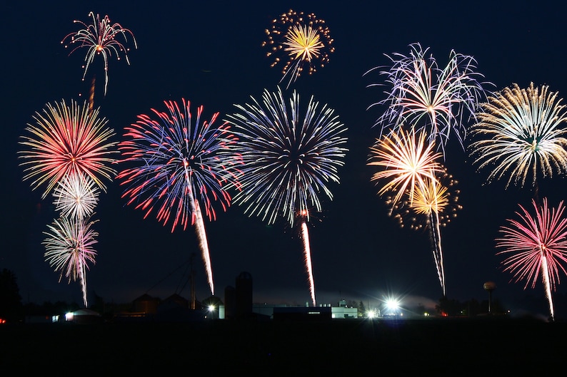 May include: A night sky filled with colorful fireworks exploding in a variety of shapes and sizes. The fireworks are set off in a field with a few buildings in the background.
