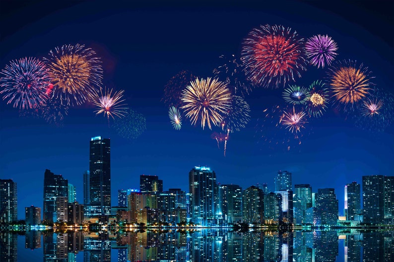 May include: A nighttime cityscape with colorful fireworks exploding in the sky. The fireworks display features red, yellow, and purple bursts above the illuminated buildings. The city lights reflect in the water below.