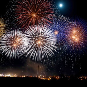 May include: A nighttime scene of colorful fireworks exploding in the sky. The fireworks display features bursts of white, gold, red, and blue against a dark background. A full moon is visible in the upper right corner, and a silhouetted town is at the bottom.
