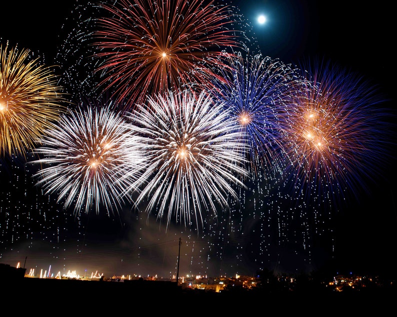 May include: A nighttime scene of colorful fireworks exploding in the sky. The fireworks display features bursts of white, gold, red, and blue against a dark background. A full moon is visible in the upper right corner, and a silhouetted town is at the bottom.