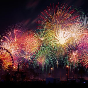 May include: A colorful fireworks display explodes in the night sky over a Ferris wheel. The fireworks are red, orange, yellow, green, and blue.