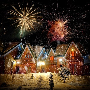 May include: A nighttime scene of fireworks exploding above a row of wooden houses covered in snow. The houses have lit windows and are decorated with string lights. Snow is falling, creating a festive winter atmosphere.