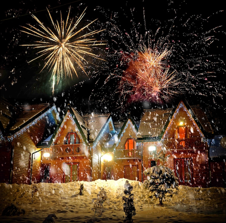 May include: A nighttime scene of fireworks exploding above a row of wooden houses covered in snow. The houses have lit windows and are decorated with string lights. Snow is falling, creating a festive winter atmosphere.