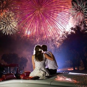 May include: A couple in a white convertible car watch a fireworks display. The fireworks are in the background and are a variety of colors, including red, white, and blue. The couple is silhouetted against the fireworks.