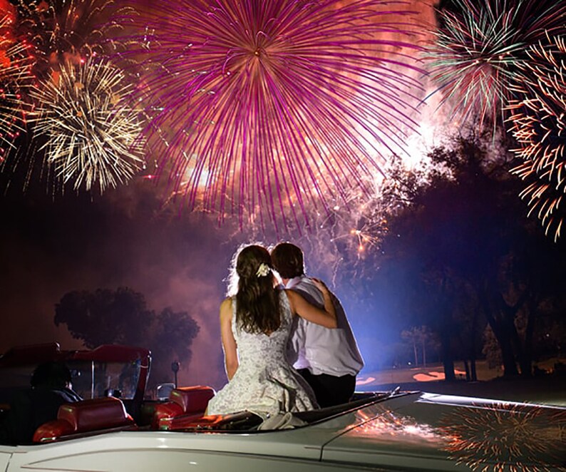May include: A couple in a white convertible car watch a fireworks display. The fireworks are in the background and are a variety of colors, including red, white, and blue. The couple is silhouetted against the fireworks.