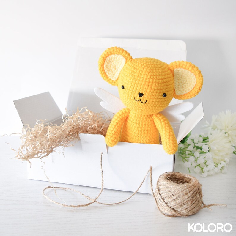 May include: A yellow crocheted stuffed animal with white wings in a white gift box with brown shredded paper. The box is tied with twine.