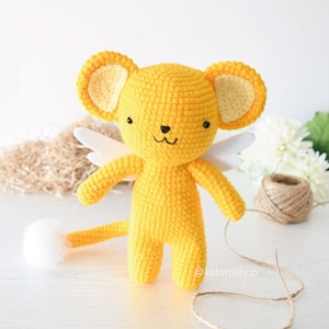 May include: A yellow crocheted stuffed animal with white wings and a tail. The animal has a cute face with black eyes and a pink nose. The animal is standing on a white surface. @koloroshp