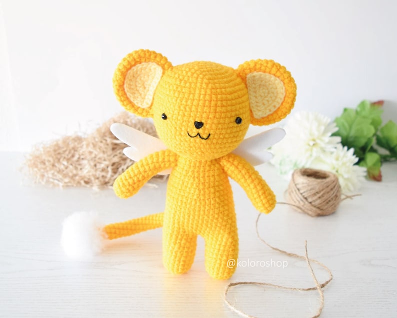 May include: A yellow crocheted stuffed animal with white wings and a tail. The animal has a cute face with black eyes and a pink nose. The animal is standing on a white surface. @koloroshp
