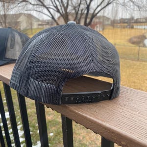 May include: Black and grey mesh trucker hat with a black adjustable strap and the word "Richardson" printed on the strap.