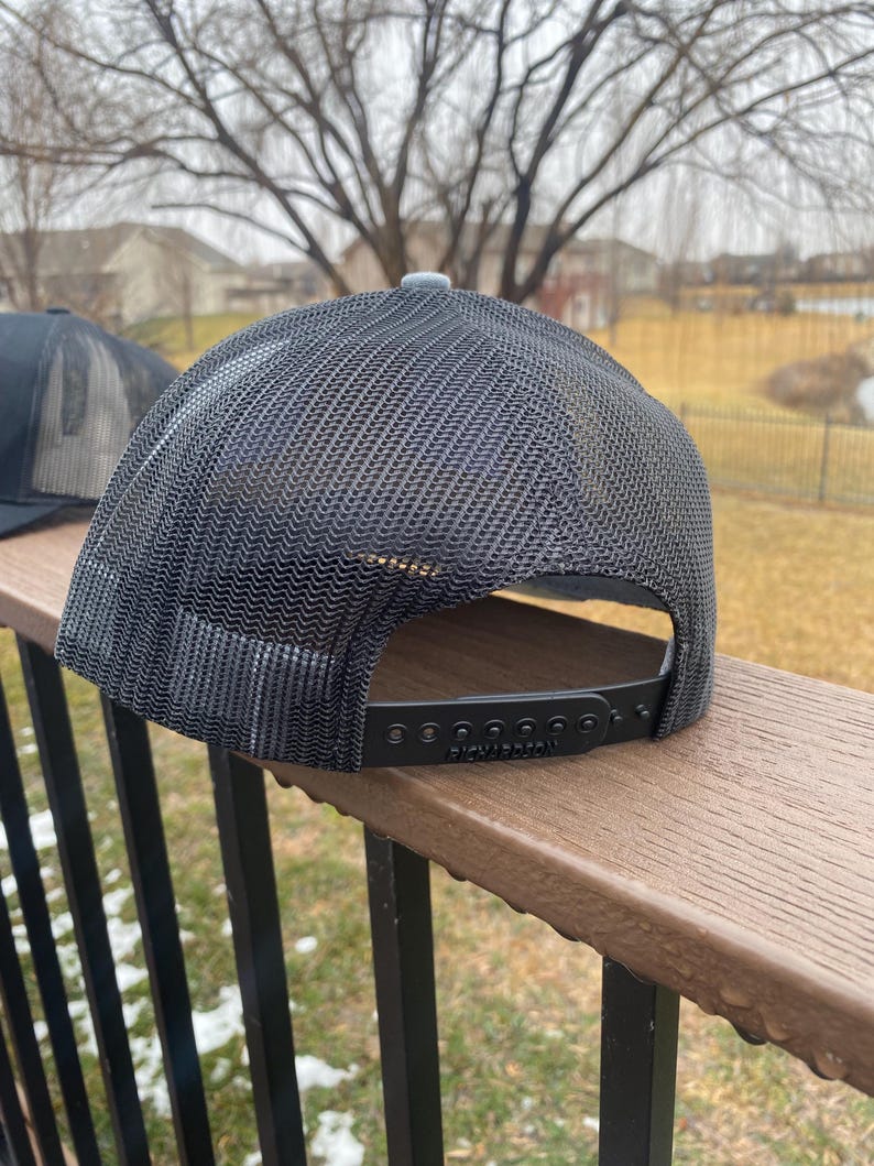 May include: Black and grey mesh trucker hat with a black adjustable strap and the word "Richardson" printed on the strap.