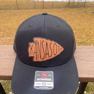 May include: Black trucker hat with a brown leather patch that says "Kansas City" in a stylised font. The hat has a mesh back and an adjustable strap with the Richardson logo and "Original 112 Trucker" text.