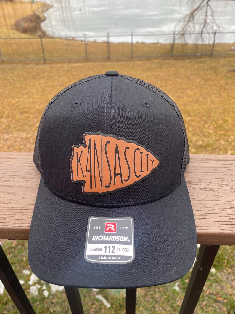 May include: Black trucker hat with a brown leather patch that says "Kansas City" in a stylised font. The hat has a mesh back and an adjustable strap with the Richardson logo and "Original 112 Trucker" text.