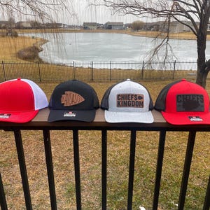 May include: Four trucker hats with different designs. The first hat is red with a white mesh back and a black logo. The second hat is black with a white mesh back and a brown leather patch with the word "Kansas" on it. The third hat is grey with a white mesh back and a brown leather patch with the words "Chiefs Kingdom" on it. The fourth hat is red with a black mesh back and a brown leather patch with the word "Chiefs" on it.