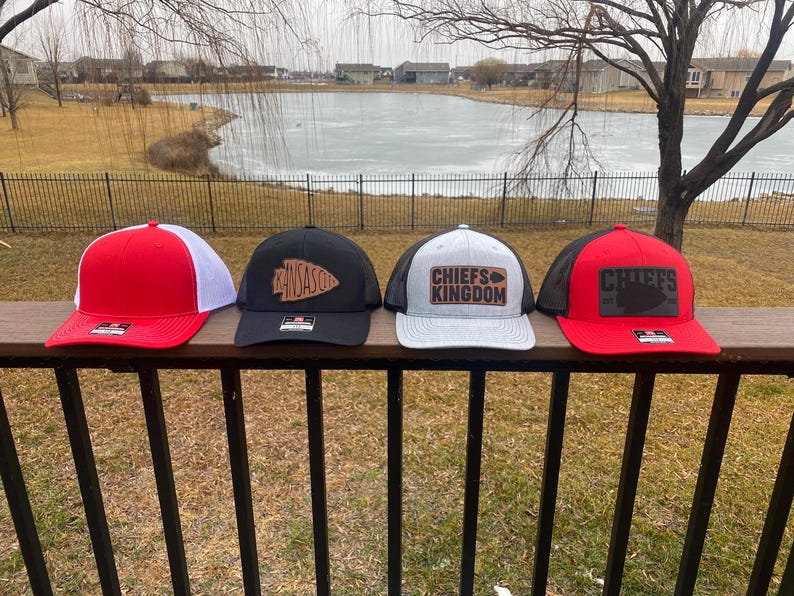 May include: Four trucker hats with different designs. The first hat is red with a white mesh back and a black logo. The second hat is black with a white mesh back and a brown leather patch with the word "Kansas" on it. The third hat is grey with a white mesh back and a brown leather patch with the words "Chiefs Kingdom" on it. The fourth hat is red with a black mesh back and a brown leather patch with the word "Chiefs" on it.
