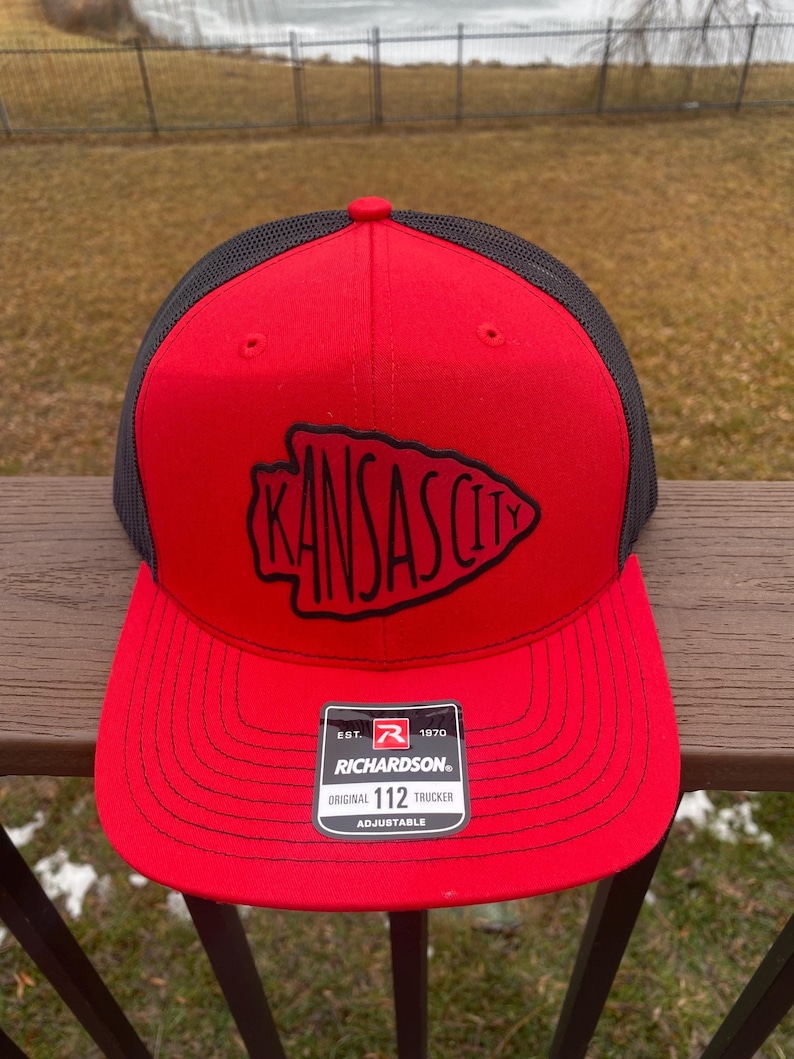 May include: Red and black trucker hat with a black embroidered Kansas City logo. The hat is adjustable and has a Richardson 112 label.