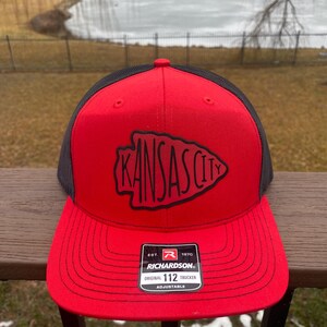 May include: Red and black trucker hat with a black embroidered Kansas City logo. The hat has a black mesh back and a red adjustable strap with a black label that reads "Richardson. Original 112 Trucker Adjustable."