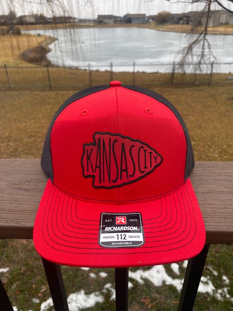 May include: Red and black trucker hat with a black embroidered Kansas City logo. The hat has a black mesh back and a red adjustable strap with a black label that reads "Richardson. Original 112 Trucker Adjustable."
