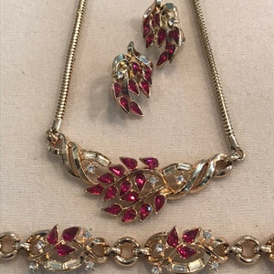 Op de afbeelding: Een gouden ketting, armband en oorbellen set met een bladontwerp. De sieraden zijn bezet met rode en heldere strass.