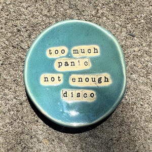 May include: A round, teal-colored ceramic trinket dish with a glossy finish. The center features the stamped black text "too much panic not enough disco" arranged in four lines, with each word individually pressed into the clay surface.