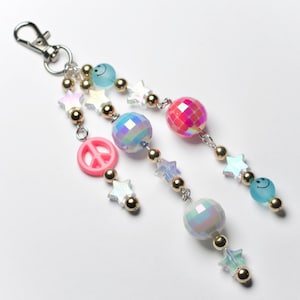 May include: A keychain with multiple dangling strands. Each strand features a mix of beads, including gold, iridescent spheres, star-shaped charms, and smiley face beads. A pink peace sign charm is also included.