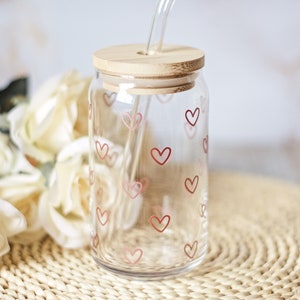 May include: A clear glass jar with a bamboo lid and a straw. The jar is decorated with a pattern of pink hearts. The jar is sitting on a woven placemat.
