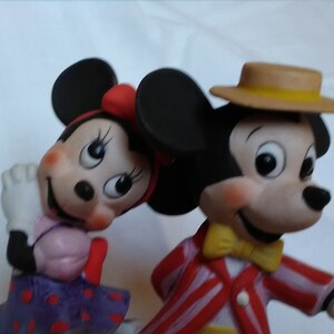 Może przedstawiać: Ceramiczna figurka Myszki Miki i Minnie Mouse. Miki ma na sobie czerwoną i białą w paski kurtkę, ż&oacute;łty krawat i brązowy kapelusz z ż&oacute;łtą wstążką. Minnie ma na sobie fioletową sukienkę w czerwone kropki i czerwoną kokardkę.