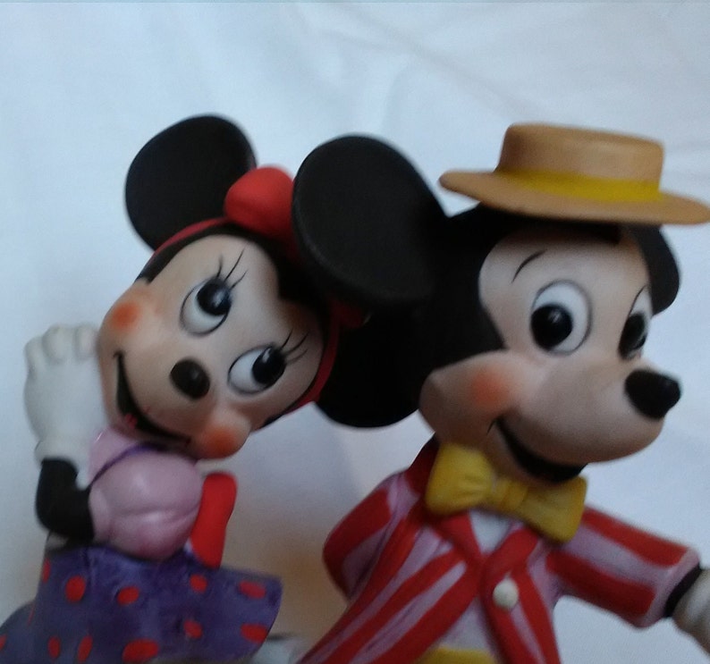 Może przedstawiać: Ceramiczna figurka Myszki Miki i Minnie Mouse. Miki ma na sobie czerwoną i białą w paski kurtkę, ż&oacute;łty krawat i brązowy kapelusz z ż&oacute;łtą wstążką. Minnie ma na sobie fioletową sukienkę w czerwone kropki i czerwoną kokardkę.