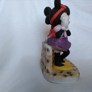 Może przedstawiać: Ceramiczna figurka Minnie Mouse w fioletowo-czerwonej sukience w kropki, z czerwoną kokardką i ż&oacute;łtymi butami. Stoi na kamiennym murze z brązowo-białym wzorem.