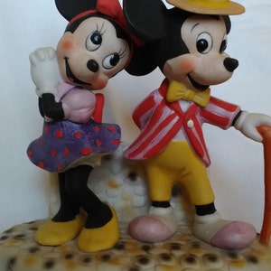 Może przedstawiać: Zestaw ceramicznych figurek Myszki Miki i Minnie. Miki ma na sobie czerwoną i białą w paski kurtkę, ż&oacute;łte spodnie, ż&oacute;łty krawat i słomkowy kapelusz. Minnie ma na sobie fioletową sukienkę w kropki z r&oacute;żową kokardką. Figurki stoją na białej podstawie z brązowym i ż&oacute;łtym wzorem.