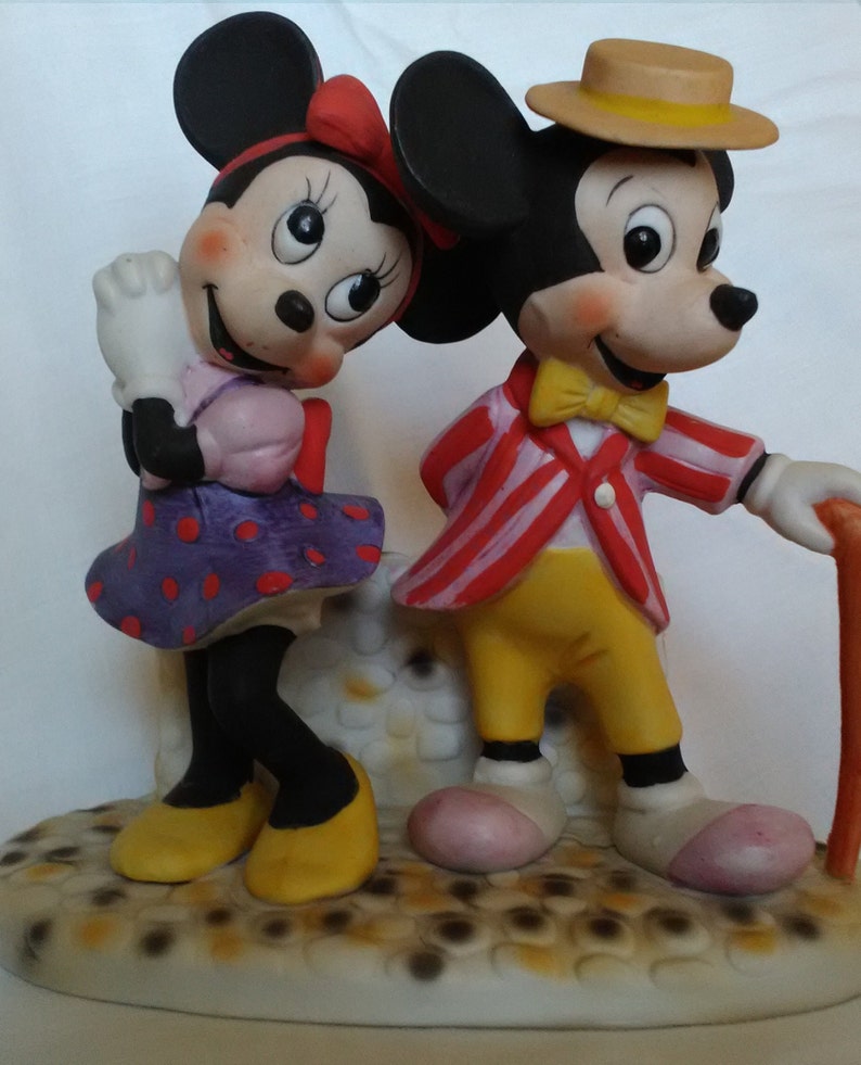 Może przedstawiać: Zestaw ceramicznych figurek Myszki Miki i Minnie. Miki ma na sobie czerwoną i białą w paski kurtkę, ż&oacute;łte spodnie, ż&oacute;łty krawat i słomkowy kapelusz. Minnie ma na sobie fioletową sukienkę w kropki z r&oacute;żową kokardką. Figurki stoją na białej podstawie z brązowym i ż&oacute;łtym wzorem.