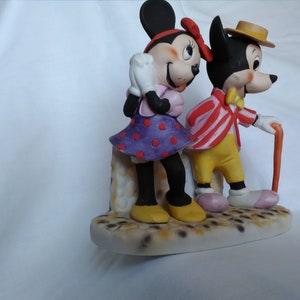 Może przedstawiać: Ceramiczna figurka Myszki Miki i Minnie Mouse. Miki ma na sobie czerwoną i białą w paski kurtkę, ż&oacute;łte spodnie i brązowy kapelusz. Minnie ma na sobie fioletową i czerwoną w kropki sukienkę. Oboje stoją na białej podstawie z brązowo-białą wzorzystą powierzchnią.