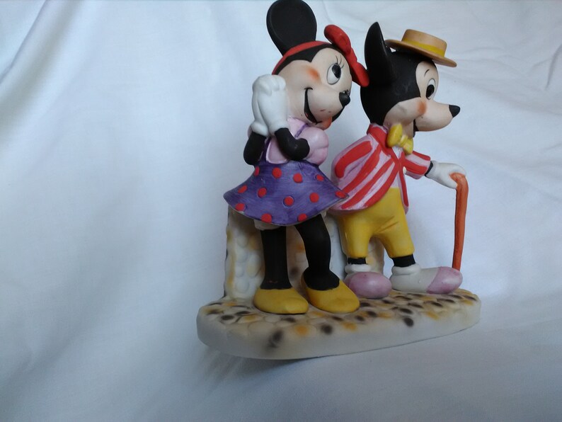 Może przedstawiać: Ceramiczna figurka Myszki Miki i Minnie Mouse. Miki ma na sobie czerwoną i białą w paski kurtkę, ż&oacute;łte spodnie i brązowy kapelusz. Minnie ma na sobie fioletową i czerwoną w kropki sukienkę. Oboje stoją na białej podstawie z brązowo-białą wzorzystą powierzchnią.