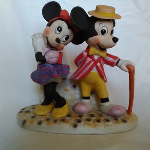 Może przedstawiać: Ceramiczna figurka Myszki Miki i Minnie Mouse. Miki ma na sobie czerwoną i białą w paski kurtkę, ż&oacute;łte spodnie i brązowy kapelusz. Minnie ma na sobie fioletową i białą w kropki sukienkę i czerwoną kokardkę. Stoją na białej podstawie z brązowo-białym wzorem.