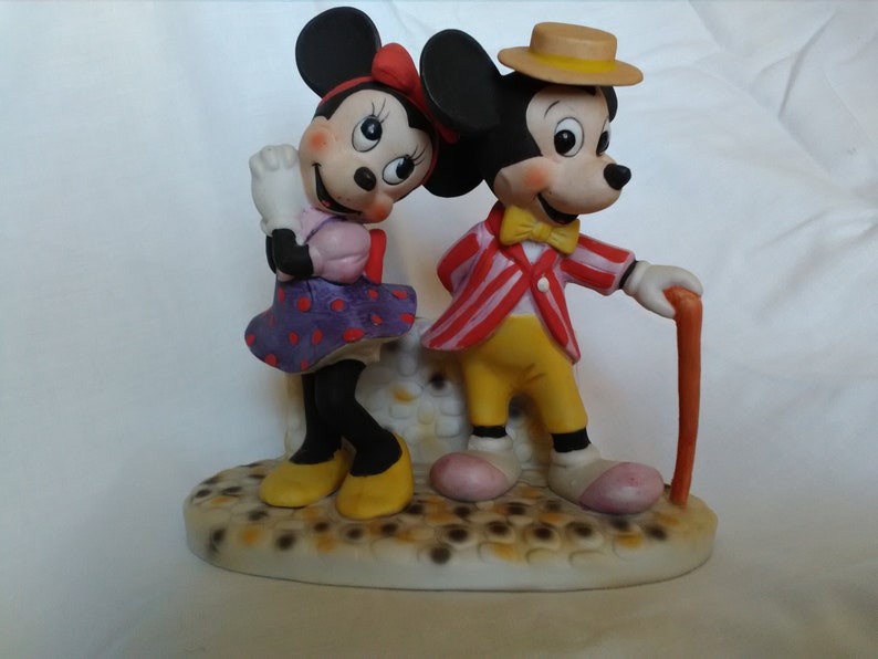 Może przedstawiać: Ceramiczna figurka Myszki Miki i Minnie Mouse. Miki ma na sobie czerwoną i białą w paski kurtkę, ż&oacute;łte spodnie i brązowy kapelusz. Minnie ma na sobie fioletową i białą w kropki sukienkę i czerwoną kokardkę. Stoją na białej podstawie z brązowo-białym wzorem.