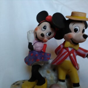 Może przedstawiać: Porcelanowa figurka Myszki Miki i Minnie Mouse. Miki ma na sobie czerwoną i białą w paski kurtkę, ż&oacute;łte spodnie, brązowy kapelusz i ż&oacute;łty krawat. Minnie ma na sobie fioletową i białą w kropki sukienkę z r&oacute;żową kokardką. Oboje się uśmiechają.