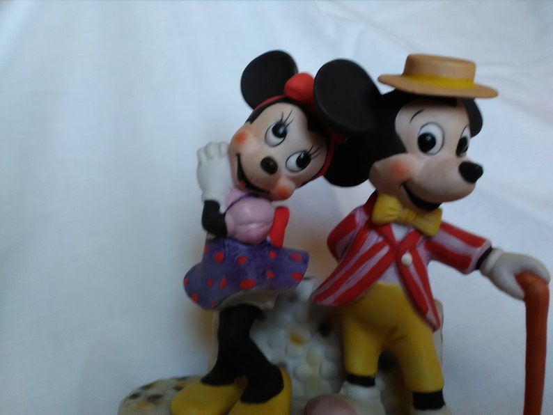Może przedstawiać: Porcelanowa figurka Myszki Miki i Minnie Mouse. Miki ma na sobie czerwoną i białą w paski kurtkę, ż&oacute;łte spodnie, brązowy kapelusz i ż&oacute;łty krawat. Minnie ma na sobie fioletową i białą w kropki sukienkę z r&oacute;żową kokardką. Oboje się uśmiechają.